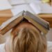 child sits at a desk with an open book over their face